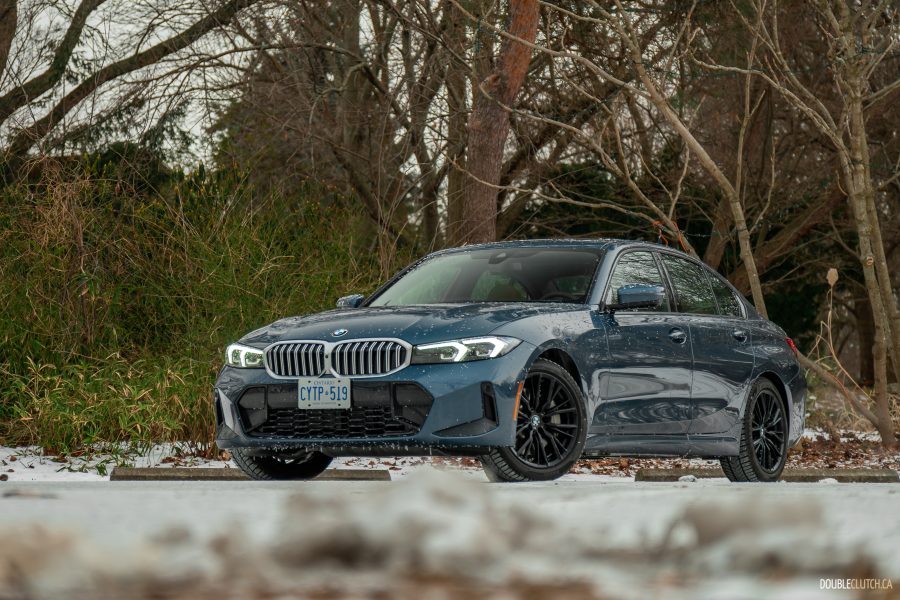 Front quarter view of a blue 2025 BMW 330i against a backdrop of trees with snow on the ground